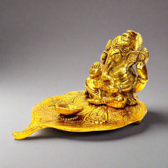 A golden metal Ganesha idol sitting on a Peepal leaf with a diya in front.
