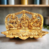 A golden metal idol of Lakshmi, Ganesha, and Saraswati with a 5-diya holder and dhupbatti stand.
