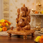 Carved wooden Ganesh idol seated on a decorative chowki with two mice, surrounded by marigold flowers and brass lamps
