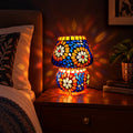 Decorative mosaic table lamp with multicolour floral glass patterns, glowing on a wooden bedside table.