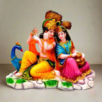 A marble dust sitting Radha Krishna idol with a peacock and a tree in the background.