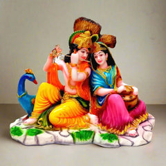 A marble dust sitting Radha Krishna idol with a peacock and a tree in the background.