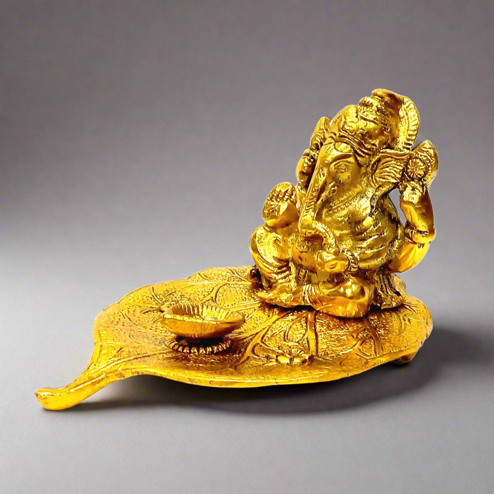 A golden metal Ganesha idol sitting on a Peepal leaf with a diya in front.