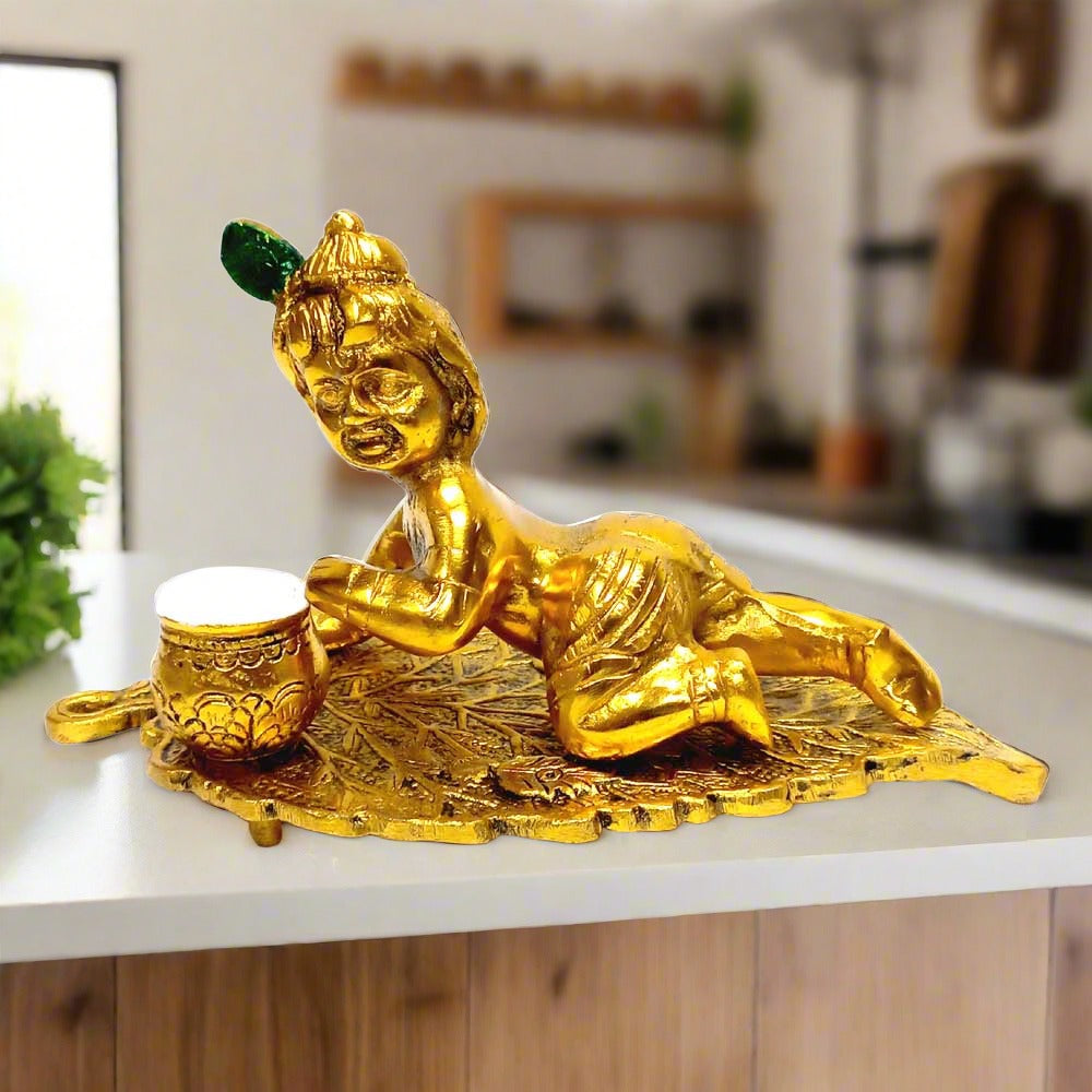 Metal Krishna idol in golden finish, seated on a leaf and eating butter.