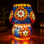 Close-up of an illuminated multicolour mosaic table lamp with blue glass tiles and glowing orange and white flowers.