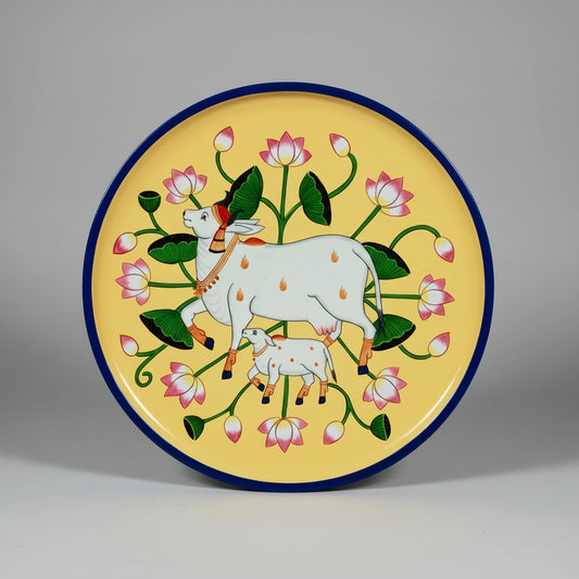 Hand-painted Pichwai art on a circular wooden wall hanging plate featuring a traditional cow and calf motif with floral borders.