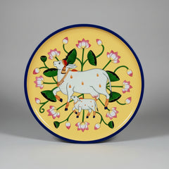 Hand-painted Pichwai art on a circular wooden wall hanging plate featuring a traditional cow and calf motif with floral borders.