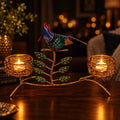 Gold-tone twin candle holder with a colourful bird, green leaves and two lit tealights on a wooden surface