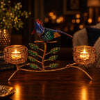 Gold-tone twin candle holder with a colourful bird, green leaves and two lit tealights on a wooden surface