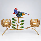 Front view of a gold-tone twin tealight holder with a colourful bird, green leaves and nest-shaped candle cups on a white background