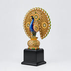 Wooden peacock memento with a blue neck, gold-toned stone-studded feathers and a decorated pedestal on a black base, shown at an angle.